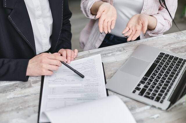 a person holding black pen pointing something on a paper discussing with someone else in front of a computer